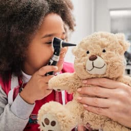 African American girl plays with her toy bear in a medical game, using an otoscope. Hearing clinic for children